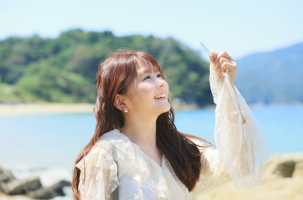 夏浜のビーチで貝殻を掲げる久間田琳加さんと、美しい海辺の風景